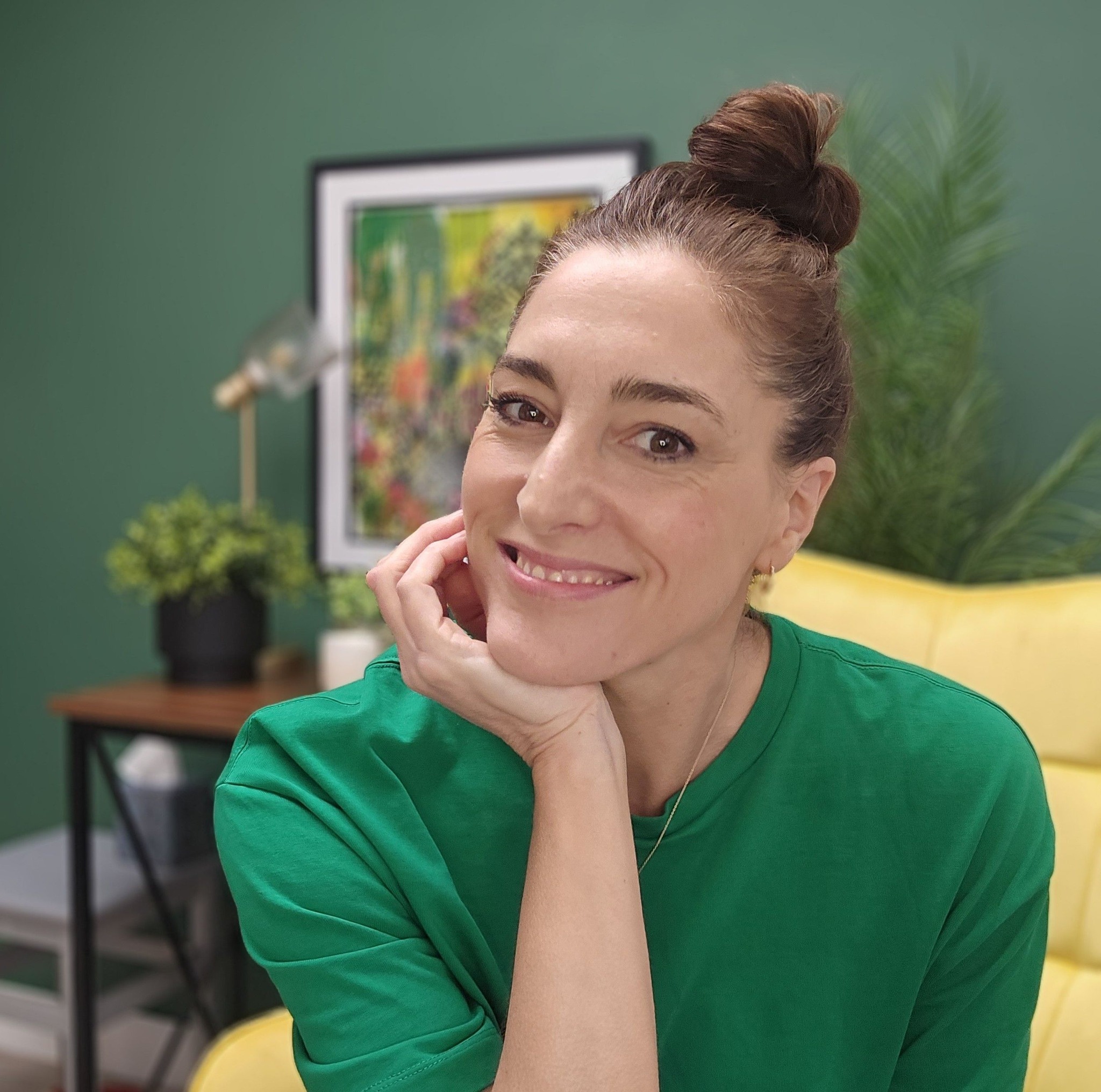 Beth Hammond Smiling sitting on a yellow chair with dark green background resting head on hand